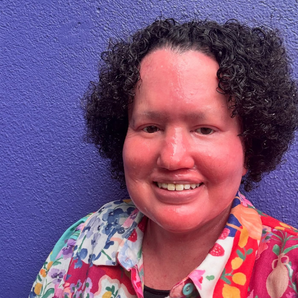 An outdoor headshot of Carly Findlay, a woman with short, dark, curly hair. She is smiling warmly at the camera, wearing a vibrant, multi-coloured button-down shirt featuring floral and strawberry patterns. She is positioned against a bright, textured purple wall that provides a bold contrast to her colourful clothing.