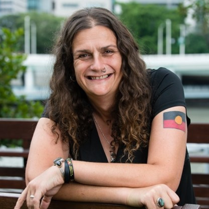 Headshot of author Claire G. Coleman with long wavy dark hair, smiling and leaning forward on a wooden railing. She is wearing a black t-shirt with an Aboriginal Flag tattoo on her arm.