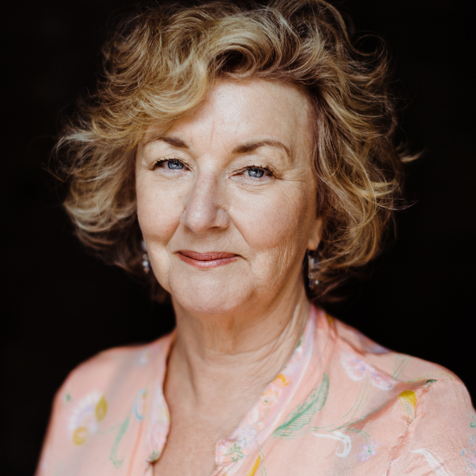 Headshot of author Debra Oswald with short, curly blonde hair and a gentle smile. She is wearing a peach-colored floral blouse against a dark, out-of-focus background.