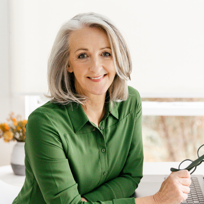 Headshot of author Joanna Nell with shoulder-length grey hair, smiling at a desk in a green shirt while holding glasses.