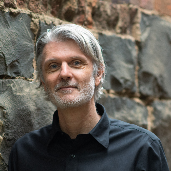 Headshot of author Rhett Davis with grey hair and a beard, wearing a black shirt and leaning against a textured stone wall.