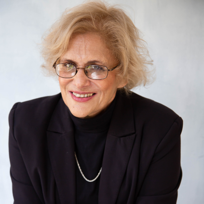 Headshot of author Susanne Gervay with short blonde hair and glasses, wearing a black blazer and turtleneck against a light background.